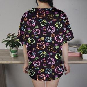May include: Black t-shirt featuring a neon-style pattern of Hello Kitty faces, flowers, stars, and suns. The design includes pink, yellow, green, and blue accents. The shirt is a loose fit and appears to be made of a soft material.
