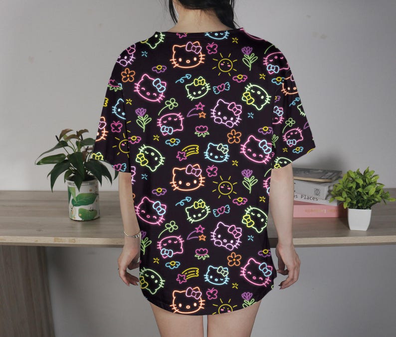 May include: Black t-shirt featuring a neon-style pattern of Hello Kitty faces, flowers, stars, and suns. The design includes pink, yellow, green, and blue accents. The shirt is a loose fit and appears to be made of a soft material.