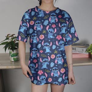Pu&ograve; includere: Una maglietta blu navy con un motivo ripetuto del personaggio Stitch di Lilo & Stitch, fiori rosa e foglie verdi. La maglietta ha maniche corte e una vestibilit&agrave; ampia.