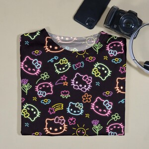 May include: Black t-shirt featuring a neon-style pattern of Hello Kitty faces, flowers, stars, and bows. The design includes pink, yellow, green, and blue accents. The shirt is folded and lies on a light beige surface.