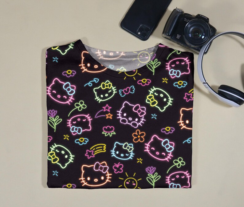 May include: Black t-shirt featuring a neon-style pattern of Hello Kitty faces, flowers, stars, and bows. The design includes pink, yellow, green, and blue accents. The shirt is folded and lies on a light beige surface.