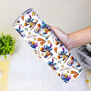 May include: A white tumbler with a repeating pattern of a blue cartoon character playing basketball. The design includes basketballs, stars, and the word "BASKETBALL". The tumbler is held in a person's hand.