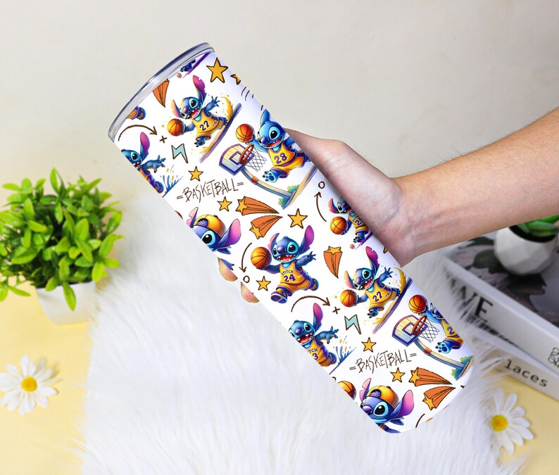 May include: A white tumbler with a repeating pattern of a blue cartoon character playing basketball. The design includes basketballs, stars, and the word "BASKETBALL". The tumbler is held in a person's hand.