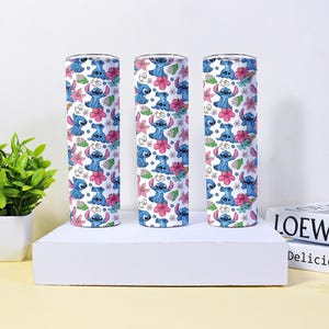 Puede incluir: Tres vasos cil&iacute;ndricos con un patr&oacute;n repetido de un personaje de dibujos animados azul, flores rosas y hojas verdes sobre un fondo blanco. Los vasos se exhiben sobre una plataforma blanca con una planta y libros en el fondo.