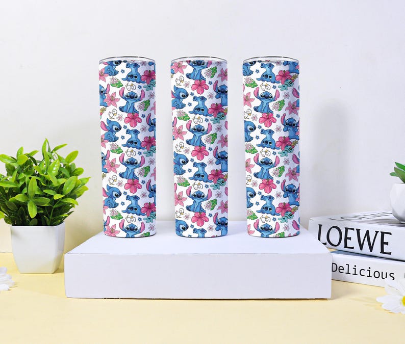 Puede incluir: Tres vasos cil&iacute;ndricos con un patr&oacute;n repetido de un personaje de dibujos animados azul, flores rosas y hojas verdes sobre un fondo blanco. Los vasos se exhiben sobre una plataforma blanca con una planta y libros en el fondo.