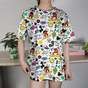 May include: White t-shirt featuring a colorful all-over print of Mickey Mouse, stars, hearts, speech bubbles, and the text "here we go!". The design includes yellow, red, black, and blue elements. The shirt has short sleeves and a round neckline.