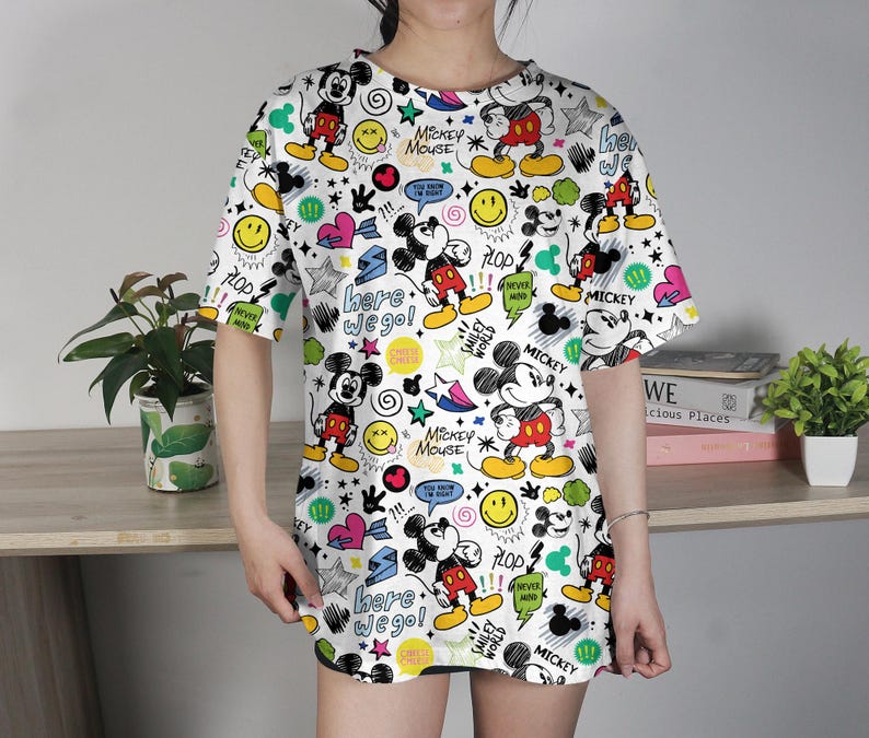 May include: White t-shirt featuring a colorful all-over print of Mickey Mouse, stars, hearts, speech bubbles, and the text "here we go!". The design includes yellow, red, black, and blue elements. The shirt has short sleeves and a round neckline.
