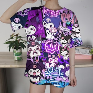 May include: A purple and black graphic t-shirt with cartoon characters and graffiti-style designs. The shirt has short sleeves and a crew neck. The design includes various characters, skulls, and text elements in white, pink, and purple.