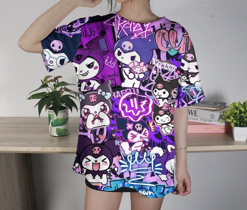 May include: A purple and black graphic t-shirt with cartoon characters and graffiti-style designs. The shirt has short sleeves and a crew neck. The design includes various characters, skulls, and text elements in white, pink, and purple.
