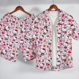Puede incluir: Un conjunto de pijama rosa con un estampado de personajes de Hello Kitty, corazones y otros gráficos. El conjunto incluye pantalones cortos y una camisa de manga corta abotonada sobre una camiseta blanca. La ropa está colgada en una percha de madera.