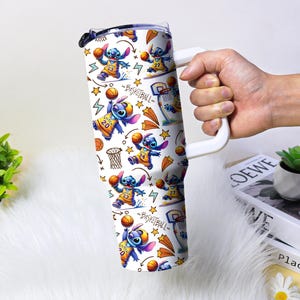 May include: A white tumbler with a handle featuring a repeating pattern of the character Stitch playing basketball. The design includes basketballs, hoops, stars, and the word "BASKETBALL". The tumbler is held by a person.
