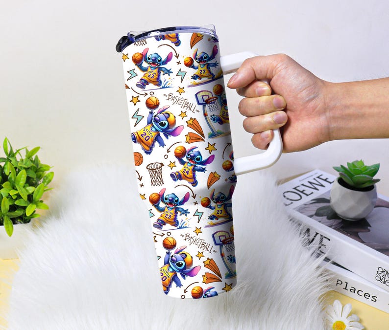 May include: A white tumbler with a handle featuring a repeating pattern of the character Stitch playing basketball. The design includes basketballs, hoops, stars, and the word "BASKETBALL". The tumbler is held by a person.
