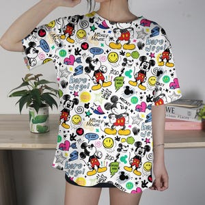May include: White t-shirt with a colorful all-over print featuring Mickey Mouse, stars, hearts, and text. The design includes the phrases "Here we go!" and "Mickey Mouse". The shirt has short sleeves.