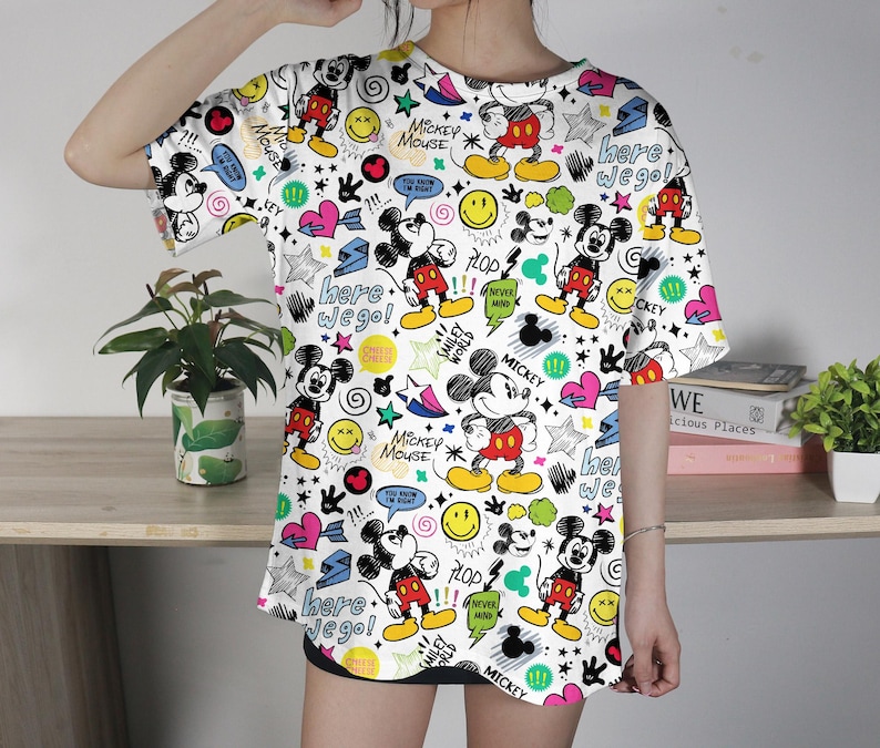 May include: White t-shirt with a colorful all-over print featuring Mickey Mouse, stars, hearts, and text. The design includes the phrases "Here we go!" and "Mickey Mouse". The shirt has short sleeves.