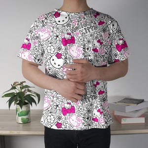 Op de afbeelding: Wit T-shirt met een herhaald patroon van Hello Kitty-personages, bloemen en de woorden "Hello Kitty" in zwart en roze. Het ontwerp is gedrukt op een shirt met korte mouwen.