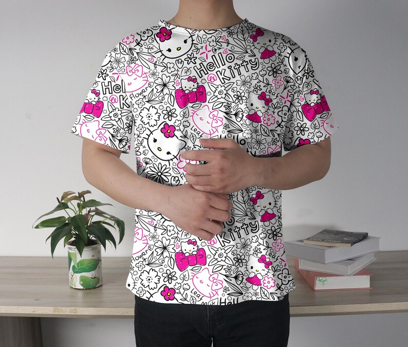 Op de afbeelding: Wit T-shirt met een herhaald patroon van Hello Kitty-personages, bloemen en de woorden "Hello Kitty" in zwart en roze. Het ontwerp is gedrukt op een shirt met korte mouwen.