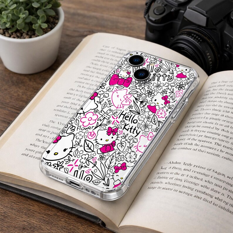 May include: Clear phone case featuring a Hello Kitty design with pink bows, flowers, and the text "Hello Kitty" on a white background. The case is on an open book with a plant and camera in the background.