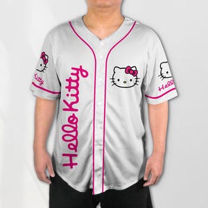 Puede incluir: Camiseta de b&eacute;isbol blanca con ribetes y detalles en rosa. Presenta el texto "Hello Kitty" en escritura rosa en la parte delantera y un gr&aacute;fico de Hello Kitty en el pecho y las mangas. La camiseta tiene una parte delantera abotonada.