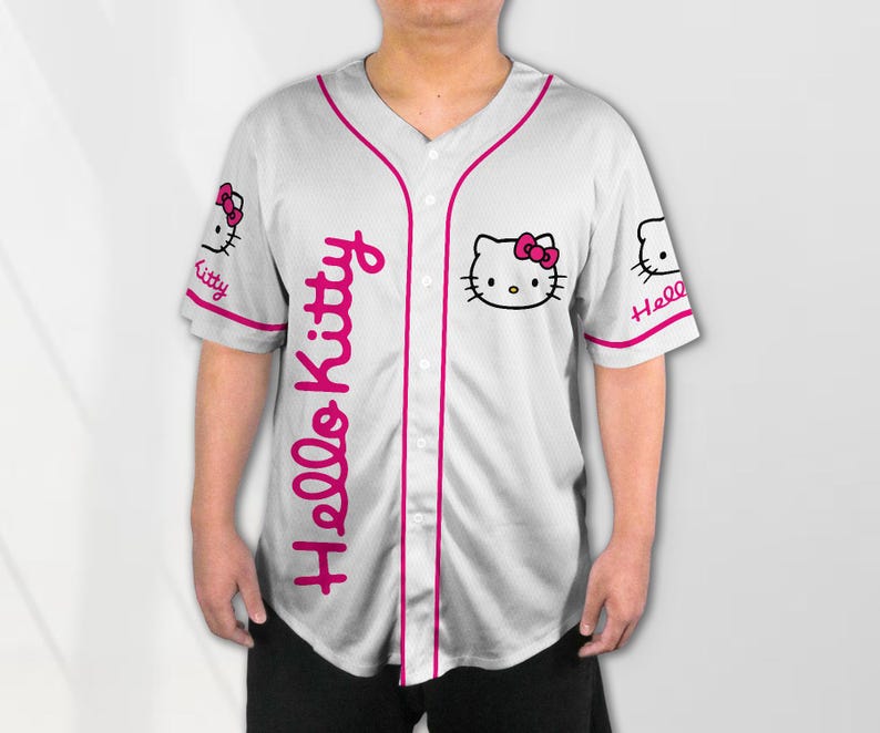 Puede incluir: Camiseta de b&eacute;isbol blanca con ribetes y detalles en rosa. Presenta el texto "Hello Kitty" en escritura rosa en la parte delantera y un gr&aacute;fico de Hello Kitty en el pecho y las mangas. La camiseta tiene una parte delantera abotonada.