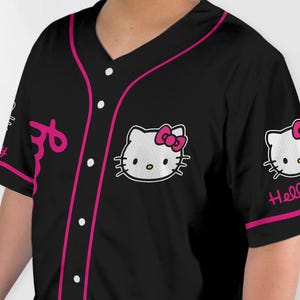 Puede incluir: Camiseta de b&eacute;isbol negra con cuello en V y mangas cortas. La camiseta presenta un ribete rosa, botones blancos y un gr&aacute;fico de Hello Kitty en el pecho y la manga. La palabra "Hello" est&aacute; escrita en la manga.