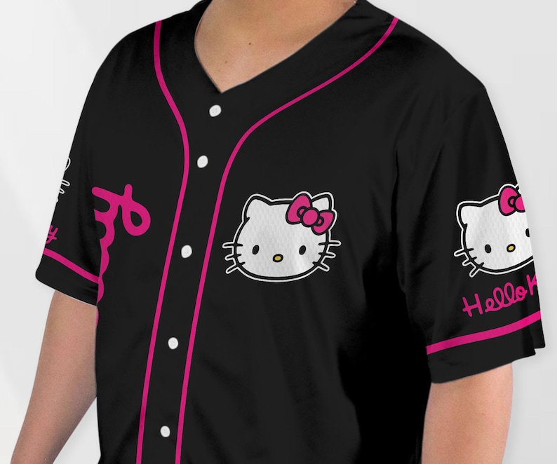 Puede incluir: Camiseta de b&eacute;isbol negra con cuello en V y mangas cortas. La camiseta presenta un ribete rosa, botones blancos y un gr&aacute;fico de Hello Kitty en el pecho y la manga. La palabra "Hello" est&aacute; escrita en la manga.