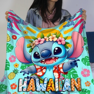 Puede incluir: Una manta azul claro con un personaje de dibujos animados con una corona de flores y una camisa hawaiana. La palabra "HAWAIIAN" se muestra en letras coloridas. La manta tiene un tema tropical con piñas y flores.