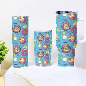 May include: Three insulated tumblers with a bright blue background and a repeating pattern featuring Homer Simpson, donuts, and sweets. The tumblers are different sizes and shapes, including a tall, a standard, and a mug.