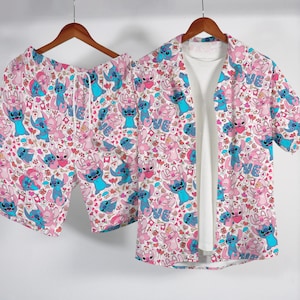 Puede incluir: Un conjunto de pijama a juego con un estampado rosa, azul y blanco de un personaje de dibujos animados y corazones. El conjunto incluye una camisa con botones, pantalones cortos y una camiseta interior blanca. La camisa y los pantalones cortos cuelgan de una percha de madera.