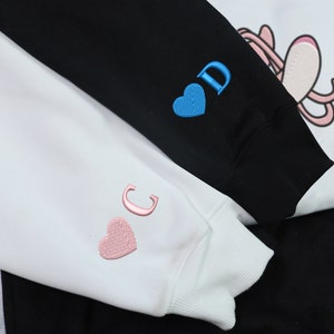 May include: Two white sweatshirts with embroidered hearts and letters. The sweatshirt on the left has a pink heart and the letter "C" embroidered on the sleeve. The sweatshirt on the right has a blue heart and the letter "D" embroidered on the sleeve.