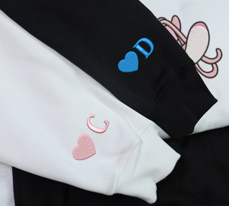 May include: Two white sweatshirts with embroidered hearts and letters. The sweatshirt on the left has a pink heart and the letter "C" embroidered on the sleeve. The sweatshirt on the right has a blue heart and the letter "D" embroidered on the sleeve.