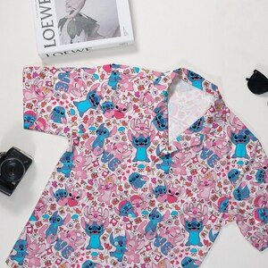 Puede incluir: Una camisa de manga corta con un estampado repetido de personajes de dibujos animados en rosa y azul sobre un fondo blanco. La camisa tiene un cuello cl&aacute;sico y est&aacute; hecha de una tela ligera. Tambi&eacute;n se ven gafas de sol y una c&aacute;mara.