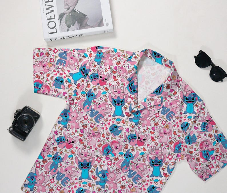 Puede incluir: Una camisa de manga corta con un estampado repetido de personajes de dibujos animados en rosa y azul sobre un fondo blanco. La camisa tiene un cuello cl&aacute;sico y est&aacute; hecha de una tela ligera. Tambi&eacute;n se ven gafas de sol y una c&aacute;mara.