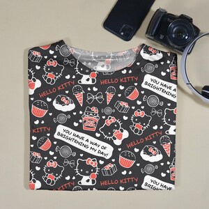May include: Black t-shirt with a repeating pattern of Hello Kitty characters, sweets, and text. The design includes red, white, and black elements. Text on the shirt reads "Hello Kitty" and "You have a way of brightening my day!"
