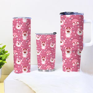 May include: Three pink tumblers with a repeating pattern of a cartoon bear, strawberries, and the text "Hi". The tumblers are different sizes, including a tall, slender one, a shorter one, and a mug with a white handle.