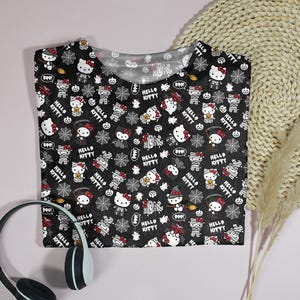 May include: Black t-shirt featuring a repeating pattern of Hello Kitty characters in Halloween costumes, spiderwebs, and the words "Hello Kitty" and "Boo!" in white. The shirt is folded on a light pink surface.