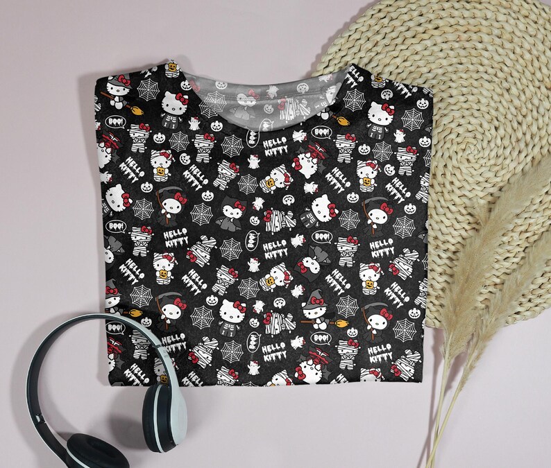 May include: Black t-shirt featuring a repeating pattern of Hello Kitty characters in Halloween costumes, spiderwebs, and the words "Hello Kitty" and "Boo!" in white. The shirt is folded on a light pink surface.