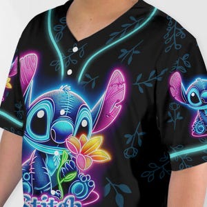 Puede incluir: Una camiseta de b&eacute;isbol negra con un gr&aacute;fico de Stitch en azul ne&oacute;n y rosa. El gr&aacute;fico muestra a Stitch sosteniendo una flor con el texto "Stitch" en la parte inferior.