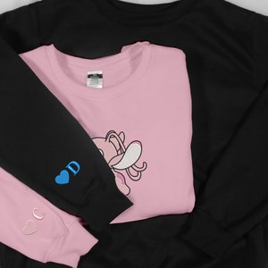 May include: Two sweatshirts, one black and one pink, are folded together. The pink sweatshirt has a cartoon character on the front and the words "C" and "D" with hearts on the sleeves.