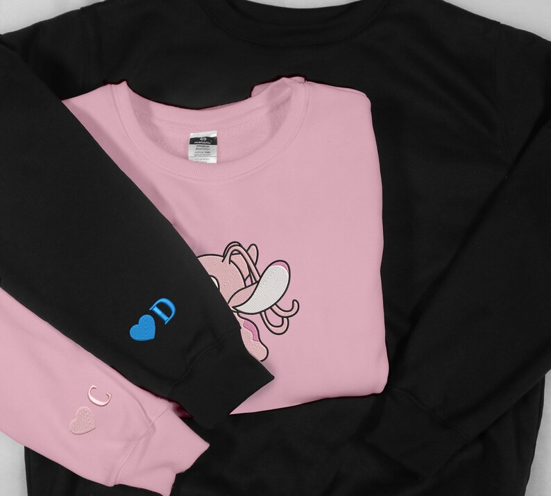May include: Two sweatshirts, one black and one pink, are folded together. The pink sweatshirt has a cartoon character on the front and the words "C" and "D" with hearts on the sleeves.