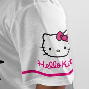 Puede incluir: Camiseta blanca con un dise&ntilde;o de Hello Kitty en la manga. El dise&ntilde;o incluye el contorno negro de la cara de Hello Kitty con un lazo rosa, y las palabras "Hello Kit" en rosa debajo de una franja rosa.
