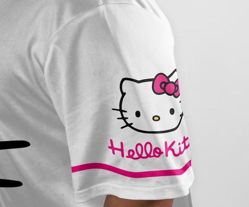 Puede incluir: Camiseta blanca con un dise&ntilde;o de Hello Kitty en la manga. El dise&ntilde;o incluye el contorno negro de la cara de Hello Kitty con un lazo rosa, y las palabras "Hello Kit" en rosa debajo de una franja rosa.