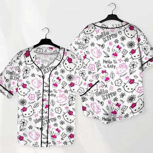 May include: White button-up shirt with a black trim and a pattern of Hello Kitty faces, flowers, and the words "Hello Kitty" in black and pink. The shirt is short-sleeved and hangs on a black hanger.