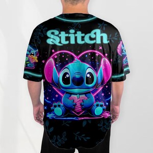 Puede incluir: Una camiseta negra con un gr&aacute;fico de Stitch de Lilo & Stitch en azul ne&oacute;n y rosa sosteniendo un coraz&oacute;n. El texto "Stitch" est&aacute; escrito en una fuente azul ne&oacute;n en la parte superior del gr&aacute;fico.