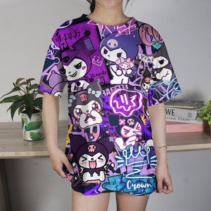 May include: Oversized purple t-shirt with a colourful all-over print of cartoon characters in different poses. The design includes graffiti art, with the word "Crown" visible. The shirt has short sleeves and a crew neck.
