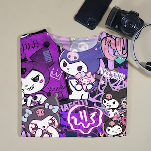 May include: A purple and white graphic t-shirt with cartoon characters and graffiti-style text. The shirt has a crew neck and short sleeves. The design includes characters with skull motifs and the word "Kuromi".