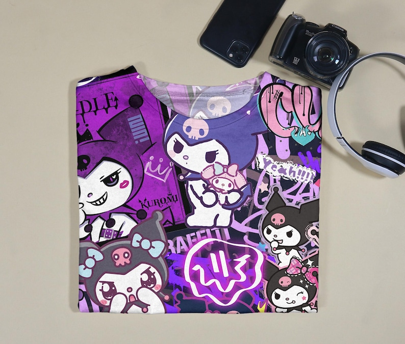 May include: A purple and white graphic t-shirt with cartoon characters and graffiti-style text. The shirt has a crew neck and short sleeves. The design includes characters with skull motifs and the word "Kuromi".