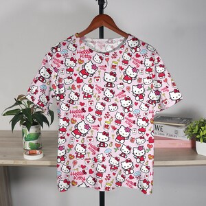 May include: A short-sleeved pink t-shirt featuring a repeating pattern of Hello Kitty characters in various poses. The design includes red bows, hearts, and the words "Hello Kitty". The shirt is hanging on a wooden hanger.
