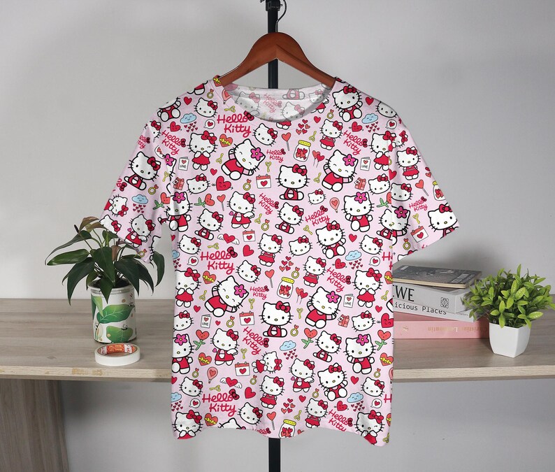 May include: A short-sleeved pink t-shirt featuring a repeating pattern of Hello Kitty characters in various poses. The design includes red bows, hearts, and the words "Hello Kitty". The shirt is hanging on a wooden hanger.