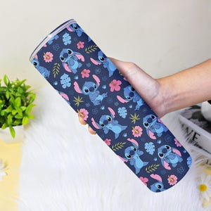 Puede incluir: Un vaso azul con un patr&oacute;n repetido del personaje Stitch de Lilo & Stitch, flores rosas y hojas verdes. El vaso se sostiene en una mano y tiene una tapa plateada.