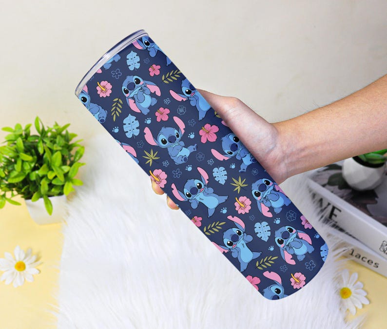 Puede incluir: Un vaso azul con un patr&oacute;n repetido del personaje Stitch de Lilo & Stitch, flores rosas y hojas verdes. El vaso se sostiene en una mano y tiene una tapa plateada.